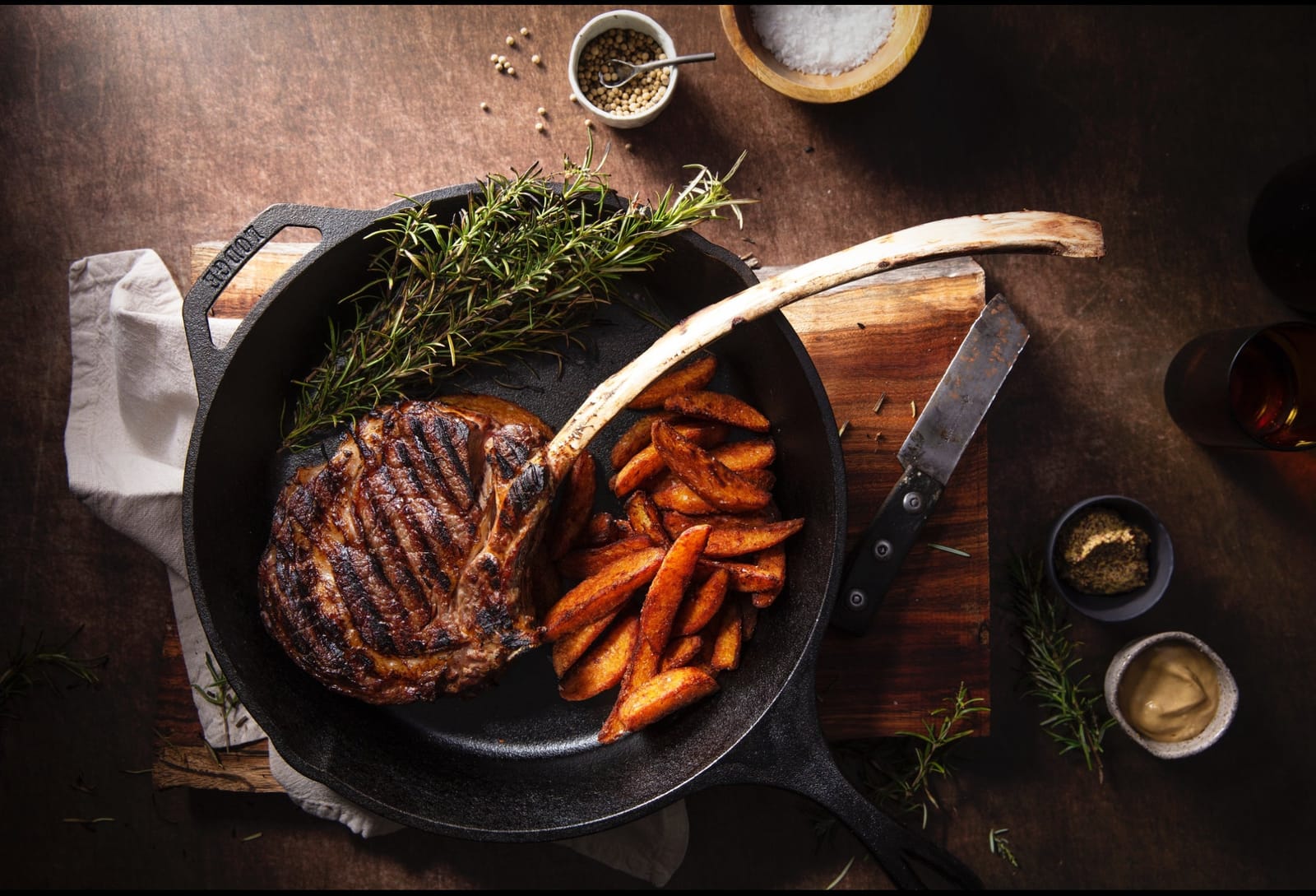 A grilled tomahawk steak resting in a cast-iron pan with roast potatoes and fresh rosemary
