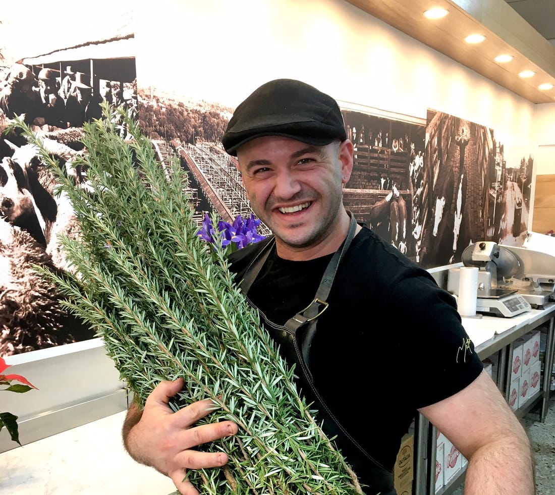 Mathew Bonnici in his flat cap and apron, holding an armful of fresh rosemary in the shop