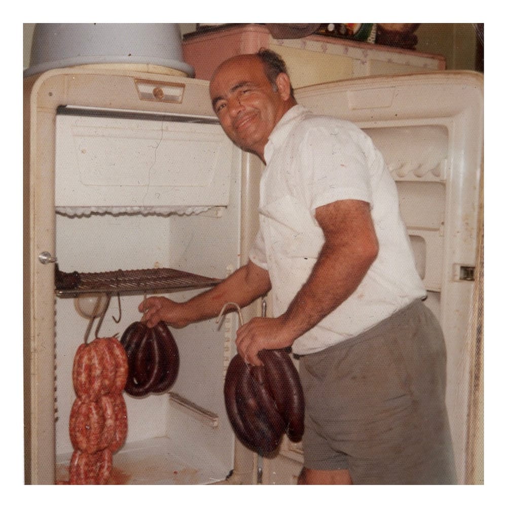 A vintage photograph of Nicolas Bonnici beside an old refrigerator hung with the family's Maltese sausages